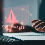 Dłoń nad klawiaturą, na ekranie ostrzeżenie "SYSTEM HACKED" i czerwony trójkąt. To znak, że system padł ofiarą złośliwego malware.