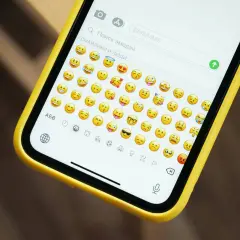 Na ekranie telefonu w żółtej obudowie widać klawiaturę z mnóstwem wesołych i smutnych emotek.