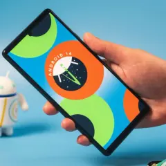 Dłoń trzyma smartfon z logo Android 14. Obok stoi figurka robota.