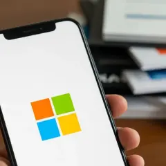 Dłoń trzyma smartfon z logo Microsoft. W tle stos dokumentów z wykresami.