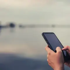 Dłonie trzymające smartfon na tle rozmytego krajobrazu.