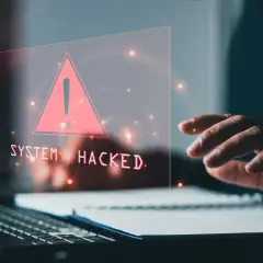 Dłoń nad klawiaturą, na ekranie ostrzeżenie "SYSTEM HACKED" i czerwony trójkąt. To znak, że system padł ofiarą złośliwego malware.