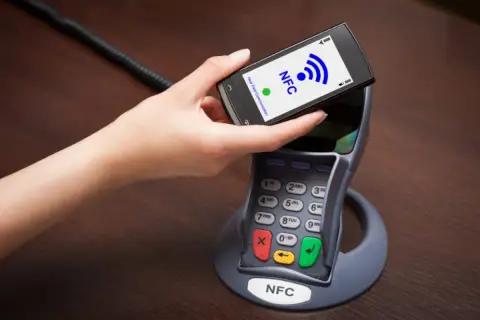 Dłoń zbliża telefon do terminala płatniczego. Na ekranie telefonu widnieje napis NFC, co oznacza technologię zbliżeniową.