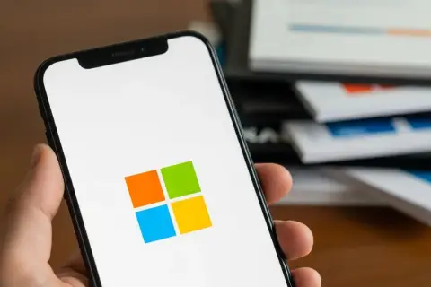 Dłoń trzyma smartfon z logo Microsoft. W tle stos dokumentów z wykresami.