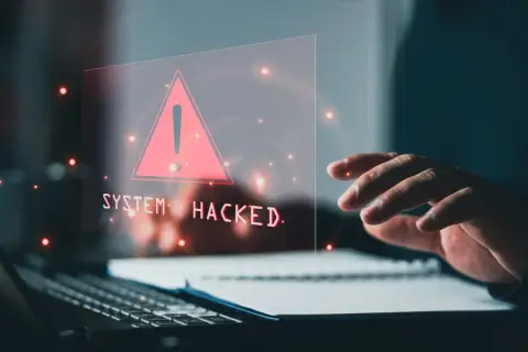 Dłoń nad klawiaturą, na ekranie ostrzeżenie "SYSTEM HACKED" i czerwony trójkąt. To znak, że system padł ofiarą złośliwego malware.