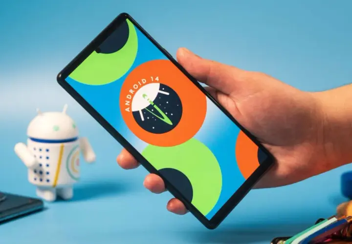 Dłoń trzyma smartfon z logo Android 14. Obok stoi figurka robota.