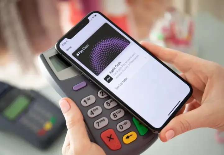 Dłoń trzyma iPhone'a nad terminalem płatniczym, ekran pokazuje "Apple Cash". To pokazuje, jak aktywować kartę i zacząć płacić.