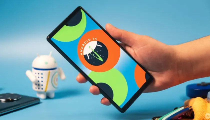Dłoń trzyma smartfon z logo Android 14. Obok stoi figurka robota.