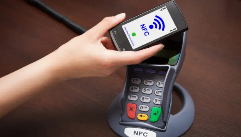 Dłoń zbliża telefon do terminala płatniczego. Na ekranie telefonu widnieje napis NFC, co oznacza technologię zbliżeniową.