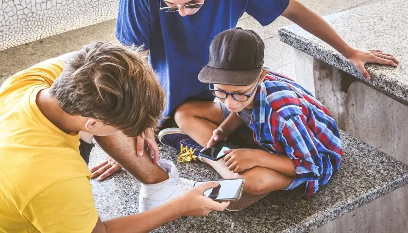 Trzech chłopców pochylonych nad telefonami. Jeden z nich pokazuje coś na ekranie, być może treści pełne hejtu, które mogą wpływać na ich samopoczucie.