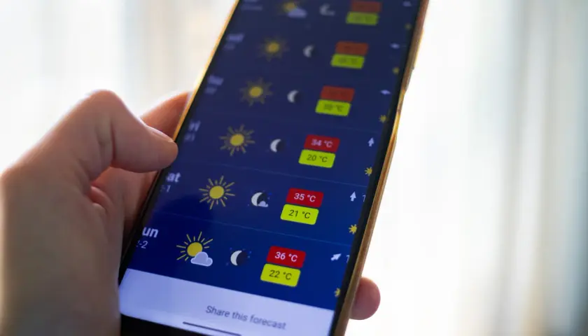 Dłoń trzyma smartfon z aplikacją pogoda, pokazującą wysokie temperatury i słoneczną aurę.