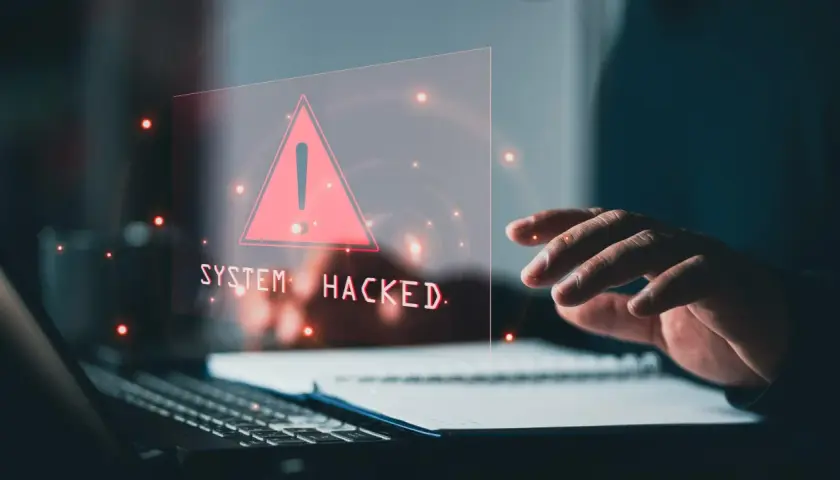 Dłoń nad klawiaturą, na ekranie ostrzeżenie "SYSTEM HACKED" i czerwony trójkąt. To znak, że system padł ofiarą złośliwego malware.