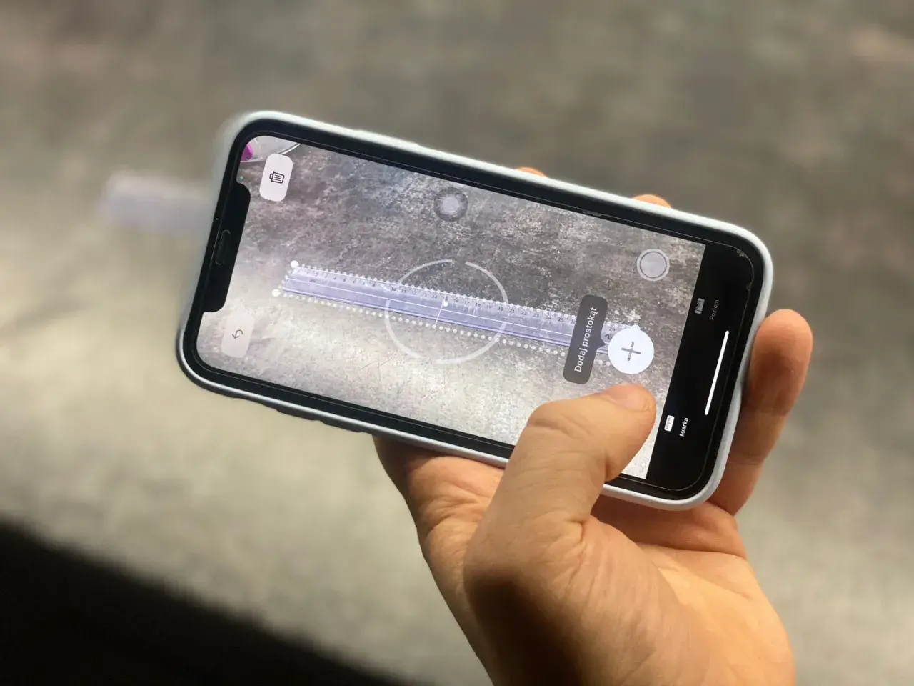 Dłoń trzyma telefon z włączoną aplikacją miarka, która mierzy odległość na powierzchni.