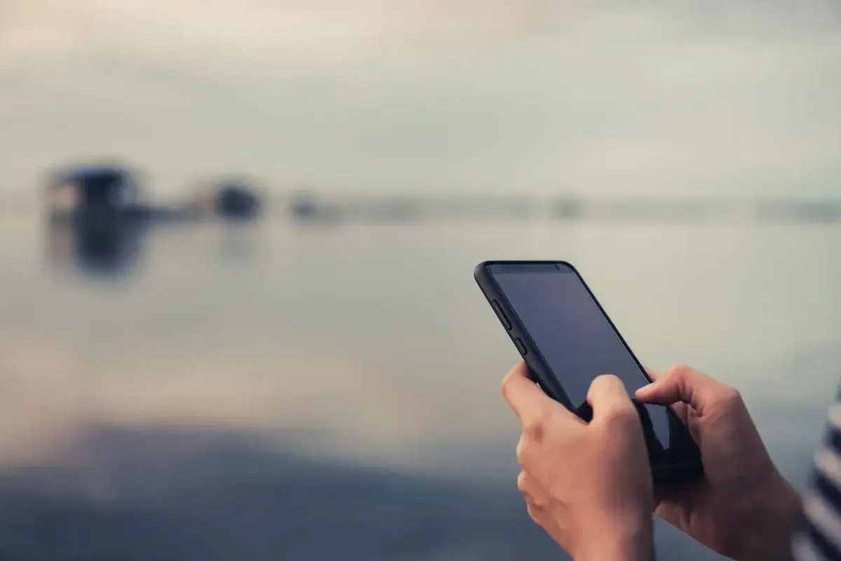 Dłonie trzymające smartfon na tle rozmytego krajobrazu.